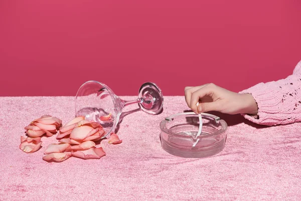 Vista ritagliata della donna che spegne la sigaretta vicino al vetro con petali sparsi sul panno di velluto isolato sul rosa, concetto girlish — Foto stock