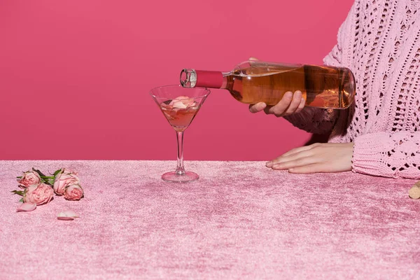 Vista cortada de mulher derramando vinho rosa perto de pétalas de rosa em pano de veludo isolado em rosa, conceito feminino — Fotografia de Stock