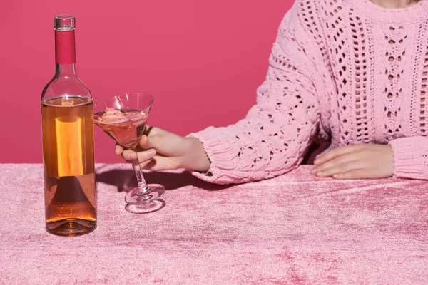 Vista recortada de la mujer sosteniendo vaso de vino rosa cerca de botella en tela de terciopelo aislado en rosa, concepto femenino - foto de stock