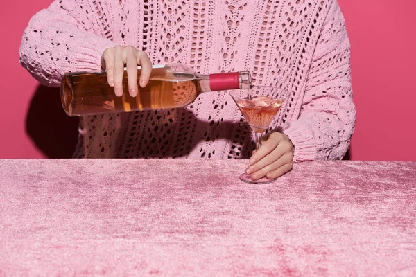 Vista recortada de la mujer vertiendo vino rosa de botella en vidrio sobre tela de terciopelo aislado en rosa, concepto femenino - foto de stock