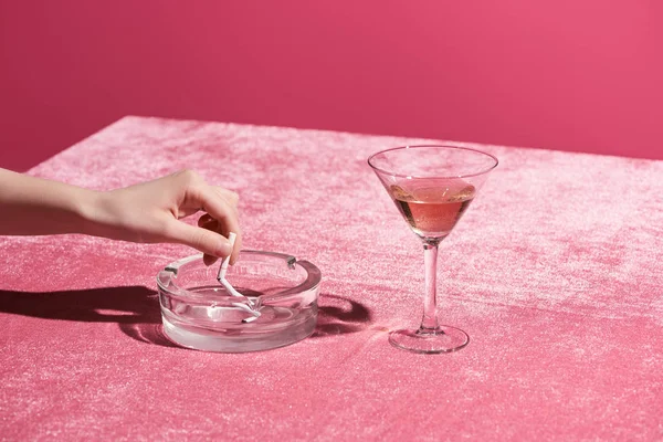 Vista cortada da mulher colocando cigarro perto de vidro de vinho rosa em pano de veludo isolado em rosa, conceito feminino — Fotografia de Stock