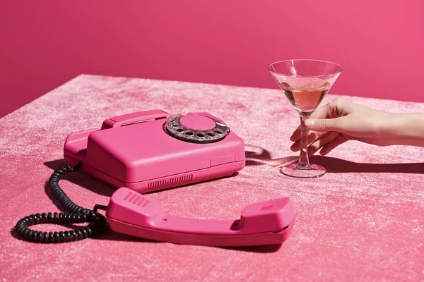 Vista cortada de mulher segurando vidro de vinho rosa perto de telefone retro em pano de veludo isolado em rosa, conceito feminino — Fotografia de Stock