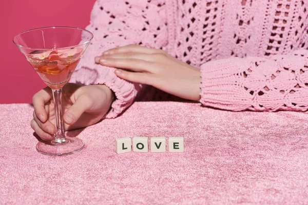 Vista cortada de mulher segurando vidro de vinho rosa perto de cubos com letras de amor em pano de veludo isolado em rosa, conceito feminino — Fotografia de Stock