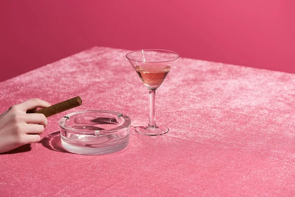 Vista cortada de mulher segurando charuto perto de cinzeiro e vidro de vinho rosa em pano de veludo isolado em rosa, conceito feminino — Fotografia de Stock