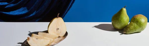 Classic still life with pear on silver plate on white table near velour curtain isolated on blue, panoramic shot — Stock Photo