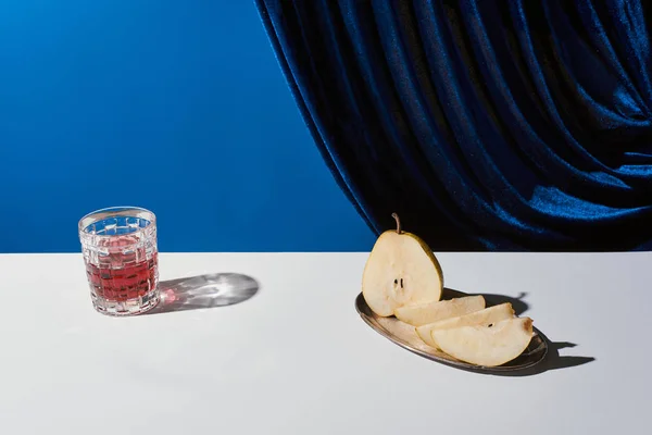 Classic still life with pear, glass with drink on white table near velour curtain isolated on blue — Stock Photo