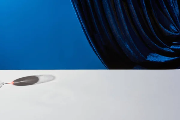 Classic still life with shadow of wine glass on white table near velour curtain isolated on blue — Stock Photo