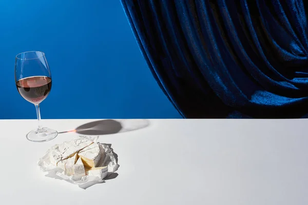 Classic still life with red wine and Camembert cheese on white table near velour curtain isolated on blue — Stock Photo