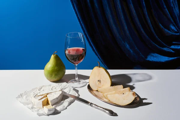 Classic still life with pears, red wine and Camembert cheese on white table near velour curtain isolated on blue — Stock Photo