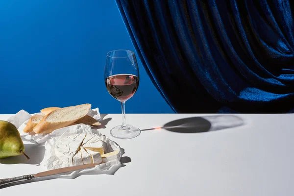 Classic still life with pear, red wine, baguette and Camembert cheese on white table near velour curtain isolated on blue — Stock Photo