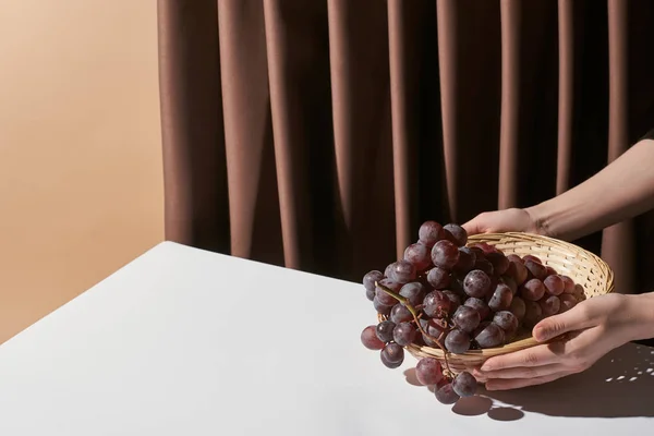 Vue recadrée de la femme tenant panier en osier avec raisin sur la table près du rideau isolé sur beige, concept de nature morte — Photo de stock