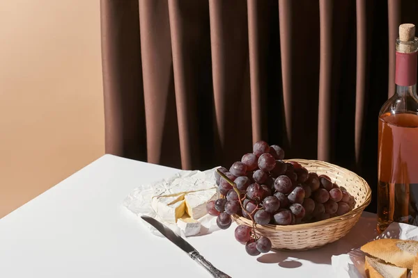 Vida tranquila clássico com vinho rosa, uva na cesta, queijo brie e baguete na mesa perto de cortina isolada em bege — Fotografia de Stock