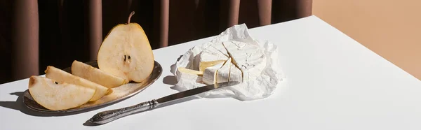 Classic still life with pear, brie cheese and knife on table near curtain isolated on beige, panoramic shot — Stock Photo