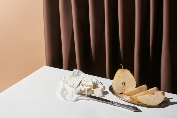 Classic still life with pear, brie cheese and knife on table near curtain isolated on beige — Stock Photo