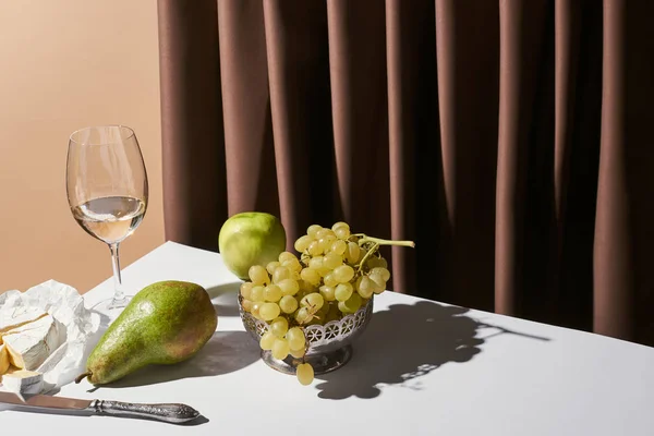 Classic still life with Camembert cheese, white wine and fruits on table near curtain isolated on beige — Stock Photo