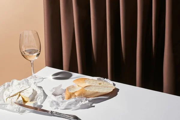 Классический натюрморт с багетом, сыр Камамбер и белое вино на столе рядом с занавесом изолированы на бежевый — стоковое фото