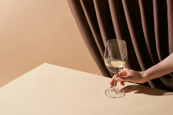 Обрізаний вид жінки, що тримає келих білого вина на столі біля завіси ізольований на бежевому, натюрморт концепції — стокове фото