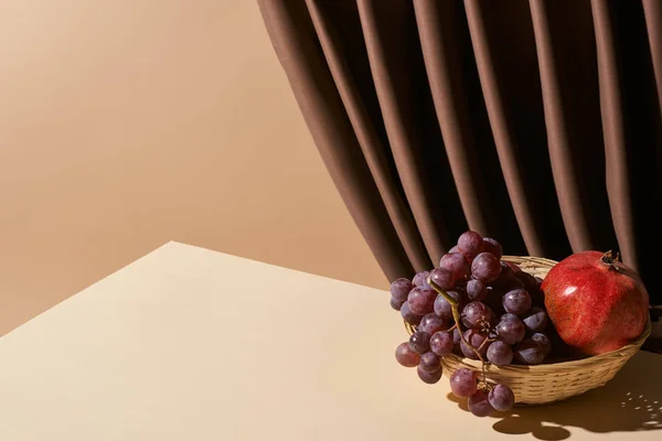 Classic still life with pomegranate and grape in wicker basket on table near curtain isolated on beige — Stock Photo