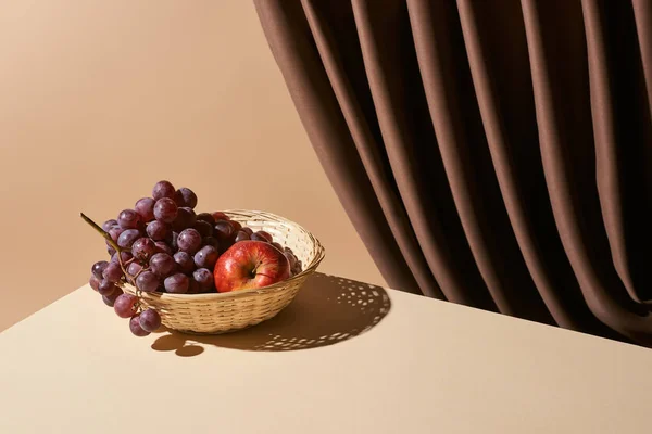 Classic still life with pomegranate and grape in wicker basket on table near curtain isolated on beige — Stock Photo