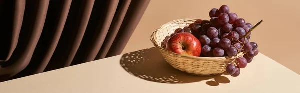 Classic still life with pomegranate and grape in wicker basket on table near curtain isolated on beige, panoramic shot — Stock Photo