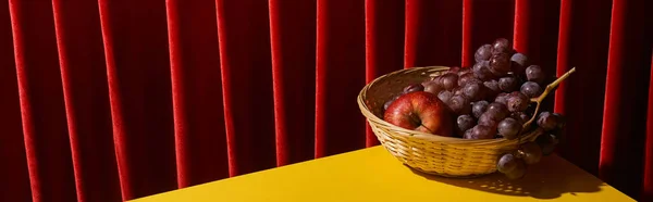 Nature morte classique avec des fruits dans le panier en osier sur la table jaune près du rideau rouge, vue panoramique — Photo de stock