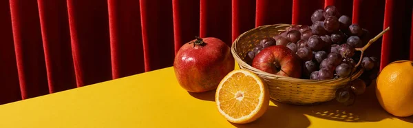 Classic still life with fruits in wicker basket on yellow table near red curtain, panoramic shot — Stock Photo