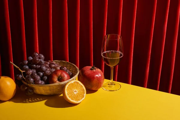 Bodegón clásico con frutas en canasta de mimbre cerca de copa de vino tinto en la mesa amarilla cerca de la cortina roja - foto de stock