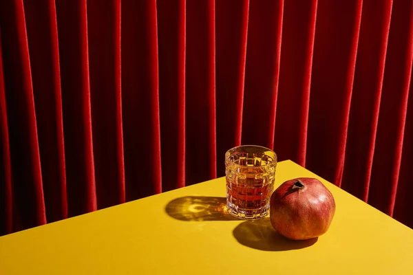 Classic still life with pomegranate near glass of red wine on yellow table near red curtain — Stock Photo