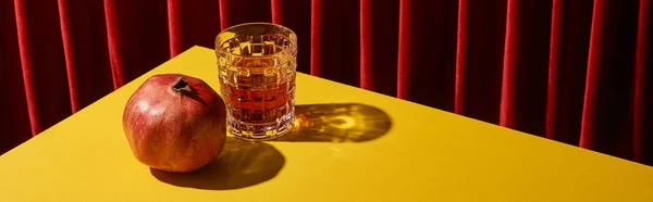 Classic still life with pomegranate near glass of red wine on yellow table near red curtain, panoramic shot — Stock Photo