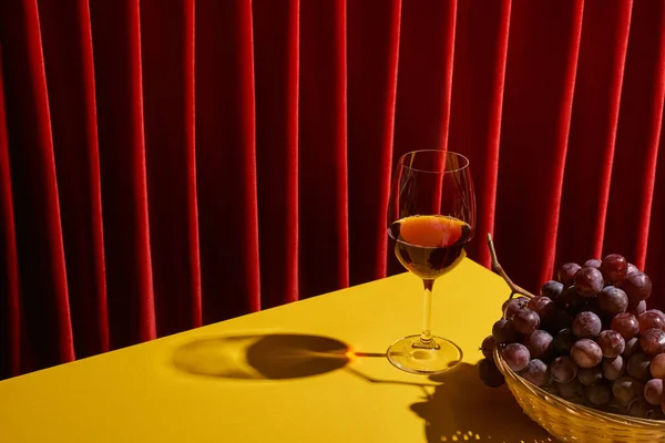 Классический натюрморт с виноградом в плетеной корзине рядом с бокалом красного вина на желтом столе возле красного занавеса — стоковое фото