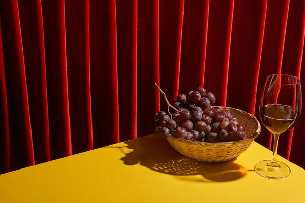 Bodegón clásico con uva en cesta de mimbre cerca de copa de vino tinto en la mesa amarilla cerca de la cortina roja - foto de stock