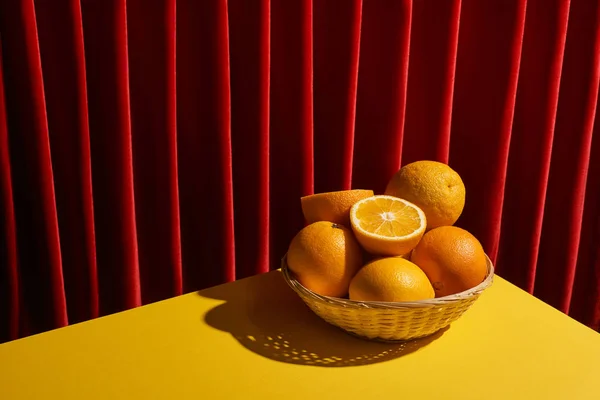 Klassisches Stillleben mit Orangen im Weidenkorb auf gelbem Tisch neben rotem Vorhang — Stockfoto