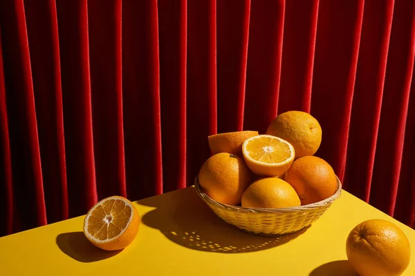 Classic still life with oranges in wicker basket on yellow table near red curtain — Stock Photo