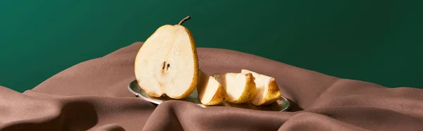 Classic still life with cut pear on silver plate on table with brown tablecloth isolated on green, panoramic shot — Stock Photo