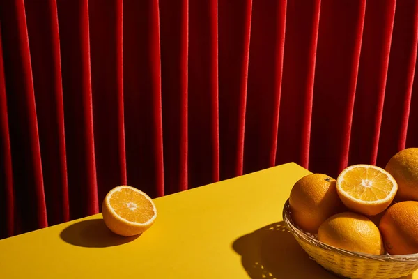 Nature morte classique avec des oranges dans le panier en osier sur la table jaune près du rideau rouge — Photo de stock