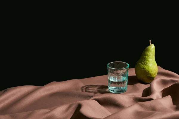 Класичне натюрморт з грушею і водою в склі на столі з коричневою скатертиною ізольовані на чорному — стокове фото
