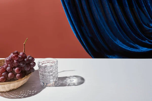 Classic still life with grape, glass on white table near velour curtain isolated on brown — Stock Photo