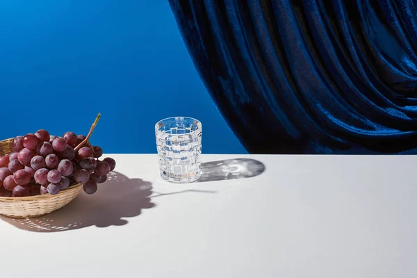 Classic still life with grape, glass on white table near velour curtain isolated on blue — Stock Photo