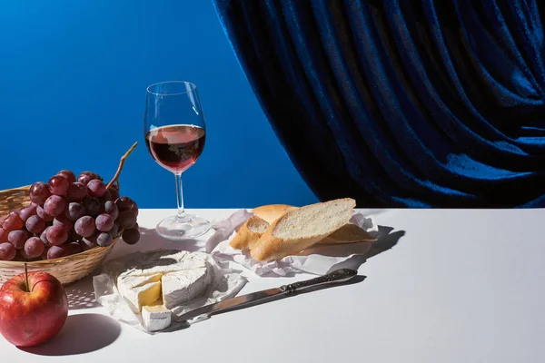 Classic still life with fruits, red wine, baguette and Camembert cheese on white table near velour curtain isolated on blue — Stock Photo