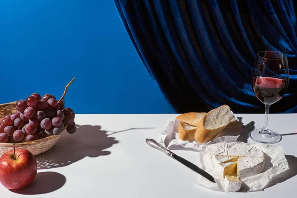 Classic still life with fruits, red wine, baguette and Camembert cheese on white table near velour curtain isolated on blue — Stock Photo
