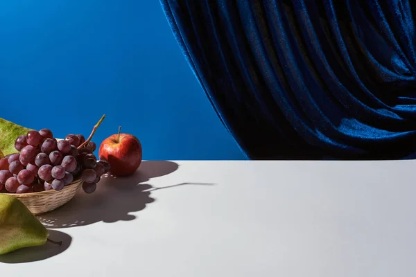 Classic still life with pears, grape and apple on white table near velour curtain isolated on blue — Stock Photo