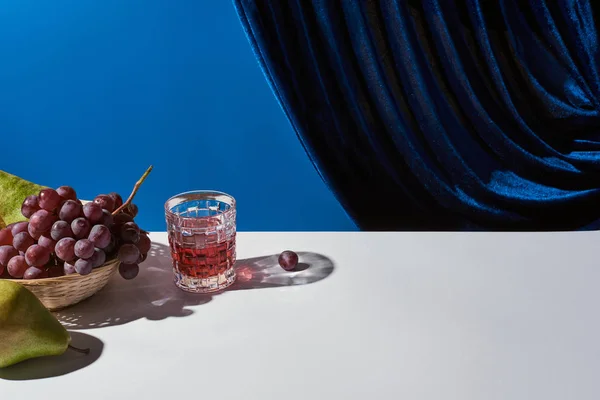 Klassisches Stillleben mit Traube, Glas mit Getränk auf weißem Tisch neben Veloursvorhang isoliert auf blau — Stockfoto