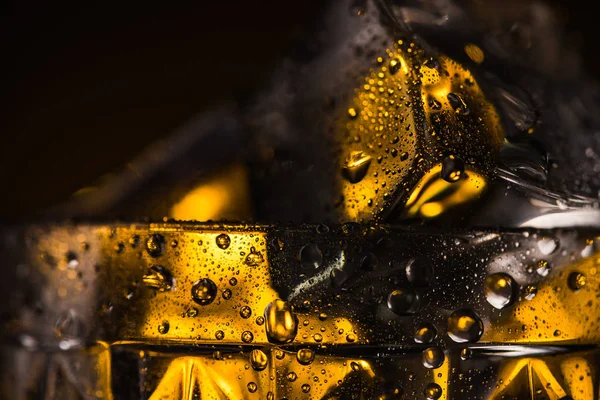 Vista de cerca del cubo de hielo cuadrado en vidrio facetado húmedo con iluminación amarilla - foto de stock