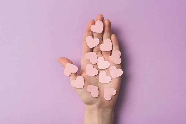 Vista recortada de la mujer sosteniendo corazones de papel rosa sobre fondo violeta - foto de stock