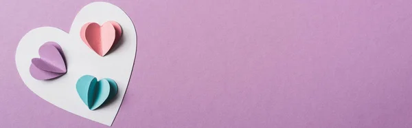 Vista superior de corazones de papel de colores en la tarjeta blanca sobre fondo violeta, plano panorámico - foto de stock