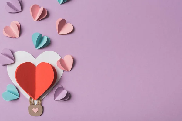 Vista superior de corazones de papel de colores alrededor de globo de aire de papel en forma de corazón sobre fondo violeta - foto de stock