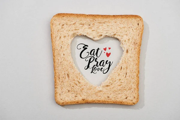 Vista superior da fatia de pão com coração esculpido e comer, rezar, amor lettering no fundo cinza — Fotografia de Stock