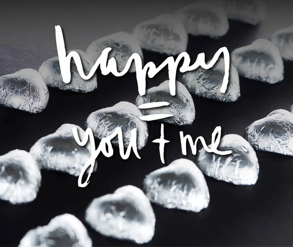 Heart shaped candies in foil on black background with happy you and me lettering — Stock Photo