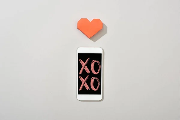 Top view of smartphone with xoxo illustration and paper heart on grey background — Stock Photo