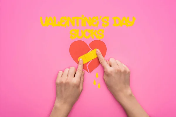 Cropped view of woman putting patch on broken paper heart isolated on pink with valentines day sucks lettering — Stock Photo